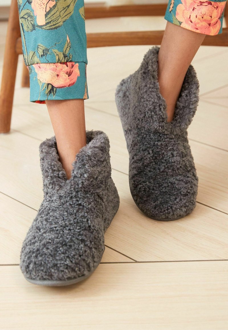 Pieds portant des chaussons gris, duveteux, montant jusqu'au milieu du mollet, posés sur un sol en bois clair, avec des jambes vêtues d'un pantalon à motifs floraux.