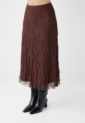 Jupe midi en dentelle marron portée avec des bottes à talons hauts noires à bout pointu sur fond blanc.