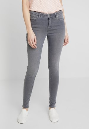 Femme portant un jean skinny gris, un haut rose clair à motifs et des chaussures blanches sans lacets, debout devant un fond uni.