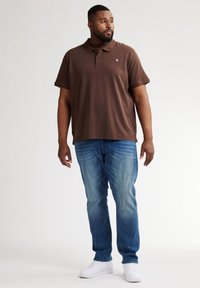 Brązowa koszulka polo z krótkim rękawem, zestawiona z jasnoniebieskimi dżinsami i białymi trampkami. Dyskretne logo na koszulce. Luźny krój.