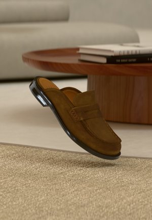 Mocasini slip-on din piele întoarsă maro plutind deasupra unei covoare bej într-un living modern cu masă de cafea din lemn și cărți stivuite.