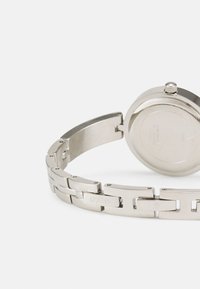 Silvrig metallklocka med länkarmband, rund urtavla och krona. Har graverat varumärkesnamn på armbandet och en slät, polerad yta.