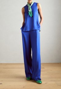 Blauwe satijnen top zonder mouwen met een afgeronde zoom, gecombineerd met bijpassende wijde broekspijpen. Een groene sjaal met patroon benadrukt de outfit. Groene schoenen maken de look compleet.