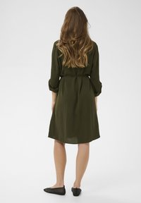 Oljčno zelena obleka do kolen z dolgimi rokavi, pasom v pasu in rahlo teksturirano tkanino. Nosi se s črnimi, ravnimi tkanimi čevlji.