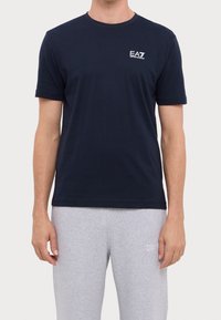 Man som bär en marinblå EA7 Emporio Armani t-shirt och ljusgråa sweatpants med texten "7 Days Active" på vänster lår.