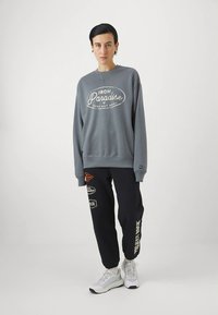 Šedá mikina s bielym logom, kombinovaná s tmavomodrými teplákmi s viacfarebnými záplatami a textom. Outfit dopĺňajú tenisky.