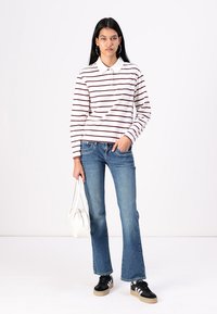 Camisa polo blanca de manga larga con rayas burdeos, combinada con jeans acampanados de color azul claro y zapatillas negras. El modelo sostiene un bolso blanco.