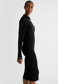 Zwarte geribbelde gebreide jurk met een kraag, lange mouwen met manchetdetails en een getailleerde pasvorm die net onder de knie valt.