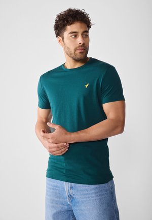 T-shirt à manches courtes teal en tissu lisse, avec un petit logo jaune sur la poitrine gauche. La coupe est décontractée et ample.