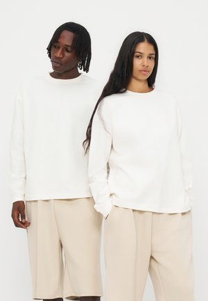 Due adulti stanno affiancati indossando magliette bianche a maniche lunghe e pantaloni beige larghi su uno sfondo bianco semplice.