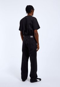 Personne avec des cheveux tressés portant un t-shirt noir, un jean noir ample avec une étiquette, et des chaussures slip-on noires et blanches, debout devant un fond uni.