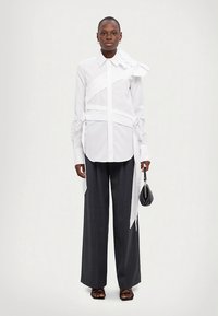 Camicia bianca con dettagli asimmetrici sulle spalline, abbinata a pantaloni scuri a gamba larga e sandali neri, tenuta con una piccola borsa nera.