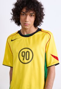 Yellow soccer jersey with short sleeves. Features black accents, a circular "90" logo, and green and red side details. Made of lightweight fabric.