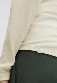 Beige geripptes Langarmshirt mit einem leichten Schwung am Saum, kombiniert mit dunkelgrünen strukturierten Hosen. Kleinere Logo-Tag in der Nähe der Taille sichtbar.