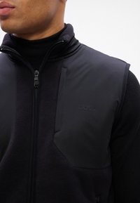 Colete preto com fecho éclair frontal, feito de tecido mate contrastante e fleece, apresentando um colarinho alto e logótipo da marca em relevo.