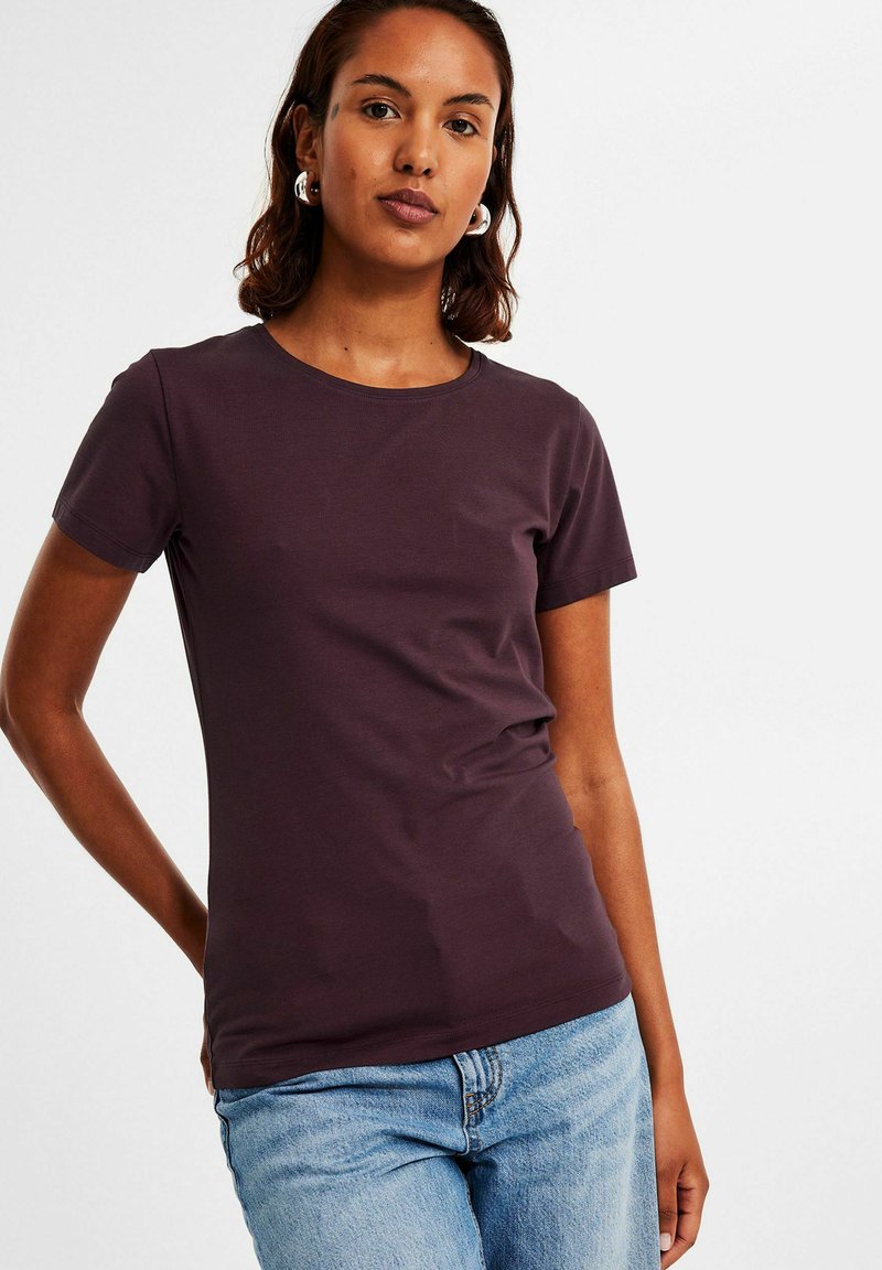 Pflaumenfarbenes T-Shirt mit kurzen Ärmeln und Rundhalsausschnitt, aus weichem Stoff, figurnaher Schnitt, kombiniert mit hellblauen Jeans.