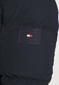 Granatowa kurtka puchowa o teksturowanym wykończeniu. Posiada prostokątną czarną naszywkę z napisem "TOMMY HILFIGER" oraz detal w czerwono-biało-niebieskich barwach.