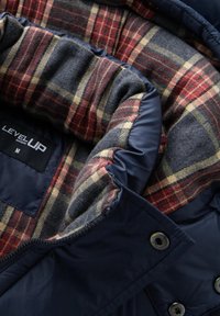 Giacca puffer blu navy con foderatura interna a quadri rossi, grigi e gialli. Presenta un'etichetta nera con la scritta "LEVEL UP" e bottoni metallici a vista.