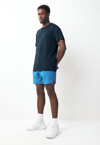 Marineblaues T-Shirt mit kurzen Ärmeln kombiniert mit hellblauen Shorts. Das Modell trägt weiße Sportschuhe und knielange weiße Socken.