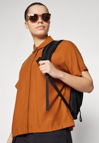 Short-sleeve, rust-colored button-up shirt with a collar; a black backpack with straps; dark sunglasses; clean background.