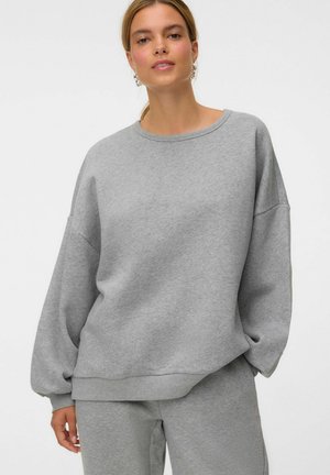Ung kvinde iført en løs grå sweatshirt og matchende joggingbukser, stående med den ene hånd i lommen mod en hvid baggrund.