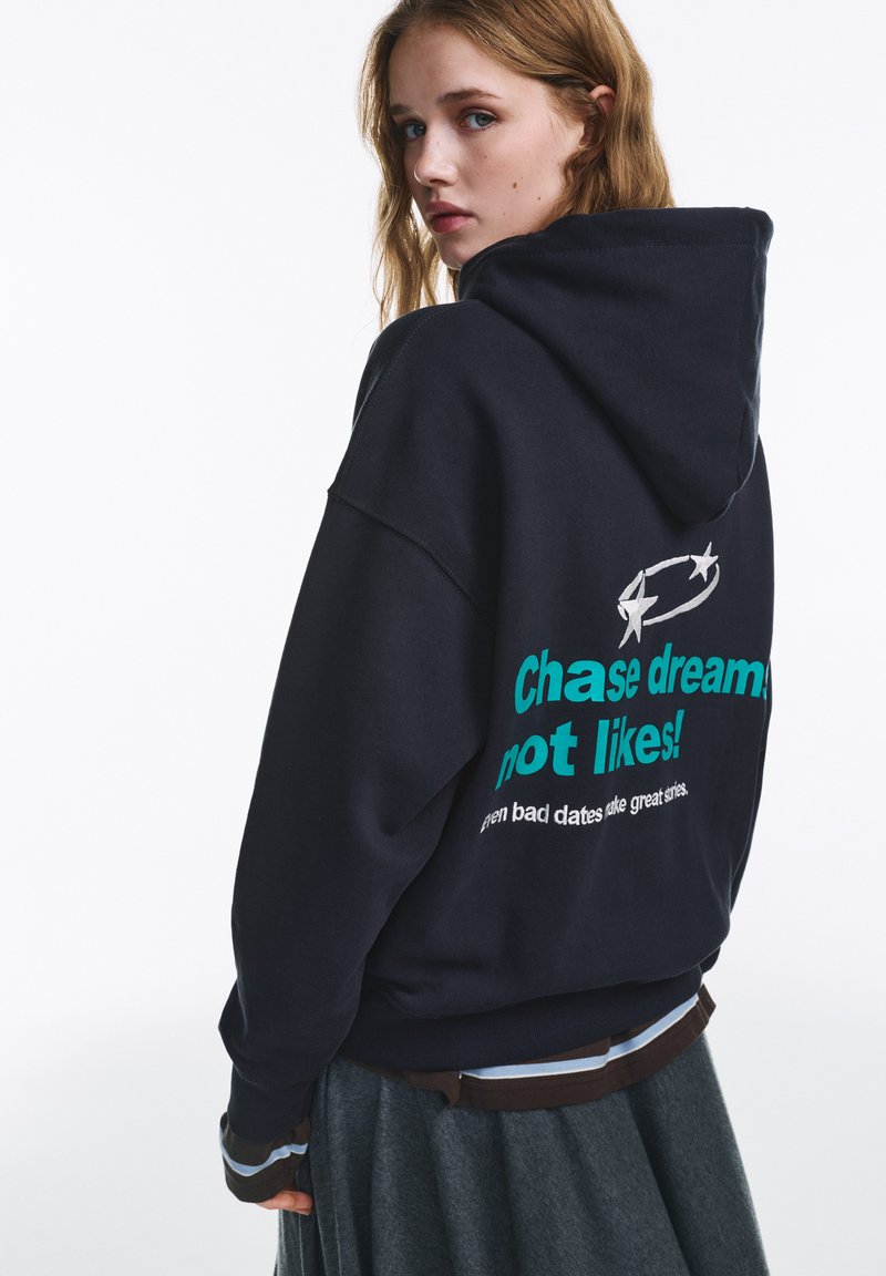 Fekete kapucnis pulóver nagy grafikával a hátulján, amely türkizkék szöveget tartalmaz "Kövesd az álmaidat, ne a lájkokat!" felirattal és alatta fehér szöveggel.