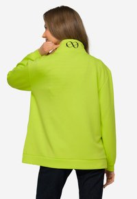 Sudadera verde neón con cuello alto, hombros caídos y un logotipo infinito negro en la parte posterior. Tela suave y ligera, corte relajado.
