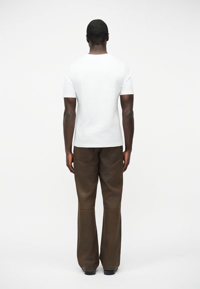 Άνδρας όρθιος πλάτη, φοράει απλό λευκό κοντομάνικο μπλουζάκι και σκούρο καφέ παντελόνι φαρδιάς γραμμής μπροστά σε λευκό φόντο.