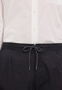 Svarta byxor med en mjuk textur och elastisk midja, med dragsko och silverfärgade aglets, bärs över en vit skjorta med knappar.