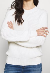 Suéter blanco de punto con cuello redondo, puños y dobladillo acanalados. La textura es de punto abierto, combinado con jeans de mezclilla azul.