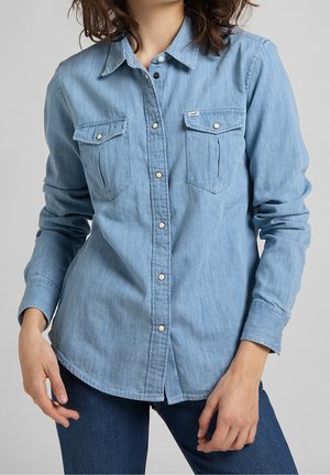 Mujer que lleva una camisa vaquera azul claro con botones de presión y dos bolsillos en el pecho, combinada con vaqueros azul oscuro.