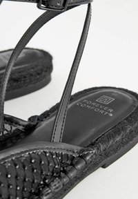 Svarta sandaler med vävda tygremmar, texturerad svart sula och en dekorativ flätad juteaccent. Har en logotyp på innersulan.