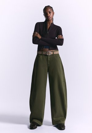 Top a coste marrone scuro con zip abbinato a pantaloni larghi olive verdi. Una cintura marrone con borchie valorizza l'outfit.