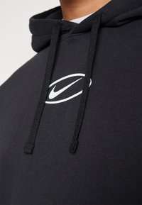 Svart munkjacka i mjukt tyg med en vit Nike-logotyp på bröstet och svarta dragsnören. Designen inkluderar en standardhuva och ribbad fåll.