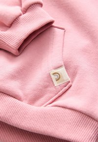 Détail de manchette et de poche de sweat-shirt rose pâle avec poignets côtelés et petite étiquette en tissu beige présentant un motif incurvé.