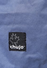 Blå texturerad tyg med en rektangulär svart lapp som har en vit lönnlöv-logotyp och texten "khujo".