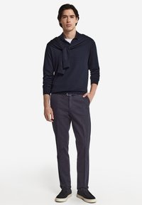 Marinblå tröja som bärs över en vit skjorta, kombinerad med mörka byxor och svarta sneakers. Snygg, figurnära design med minimala detaljer.