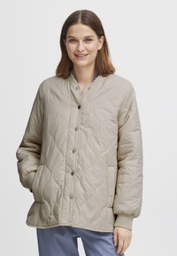 Beige quiltad jacka med rund hals, tryckknappar, sidofickor och ribbade ärmar. Slät, lättviktsmaterial med en smidig textur.