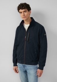 Marineblauwe ritsjack met hoge kraag, twee voorzakken en elastische boorden. Heeft een contrasterende binnenvoering en logo-detail op de mouw.