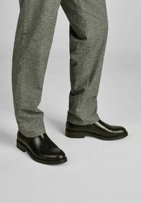 Crne kožne čizme s glatkom površinom, blagom petom i ušivenim detaljima, u paru sa sivim hlačama od teksturiranog materijala.