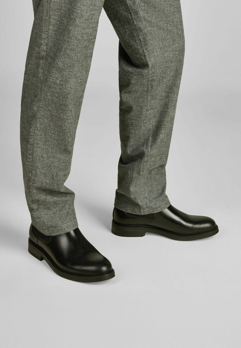 Crne kožne čizme s glatkom površinom, blagom petom i ušivenim detaljima, u paru sa sivim hlačama od teksturiranog materijala.