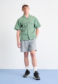 Grön kortärmad skjorta med två bröstfickor, ihopparad med grå shorts och svarta sneakers, avslutat med en beige keps.