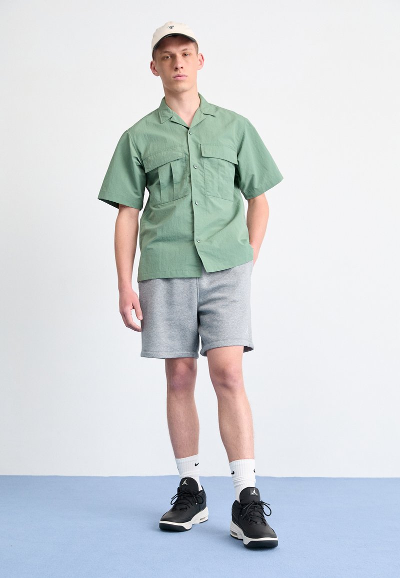 Camisa verde de manga corta con dos bolsillos en el pecho, acompañada de pantalones cortos grises y zapatillas negras, rematada con una gorra beige.