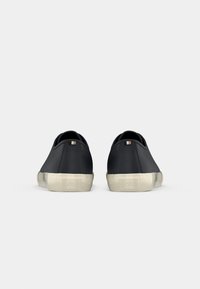Paire de baskets basses noires vues de dos avec des semelles en caoutchouc écru et de petites languettes en tissu sur fond gris.