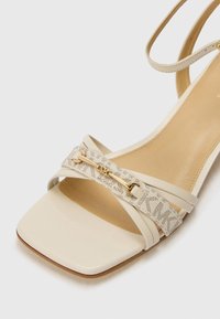 Sandale beige à bout ouvert avec bride à la cheville, bout carré et bride avant en tissu avec garnitures dorées et marque Michael Kors.
