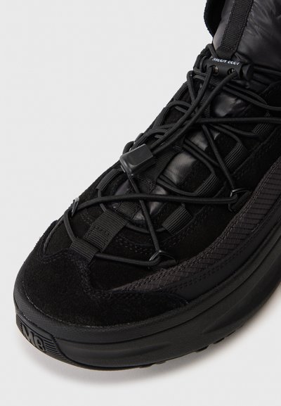 Primo piano di una sneaker nera con punta e suola testurizzata, tomaia in camoscio e tessuto, e lacci elastici bungee con fermo in plastica.