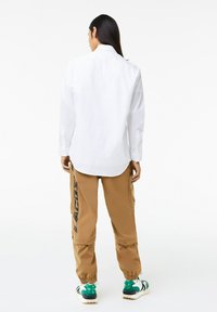 Witte langemouwen shirt met knoopmanchetten, gecombineerd met tan cargobroek met een zijprint en elastische enkelmanchetten, gedragen met groene sneakers.