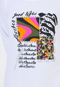 Weißes Langarmhemd mit einem Patchwork-Design und auffälligen Mustern: Pink, Orange, Grün, Grau und Tarnmuster mit dekorativen Textakzenten.