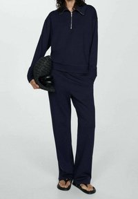 Granatowy sweter z zamkiem i długimi rękawami, w zestawie z szerokimi spodniami. Uzupełniony klapkami i trzymany w dłoni teksturowany czarny kapelusz.