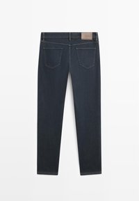 Donkerblauwe spijkerbroek gezien van achteren, met twee achterzakken, riemlussen en een cognackleurig leren merkembleem op de tailleband.